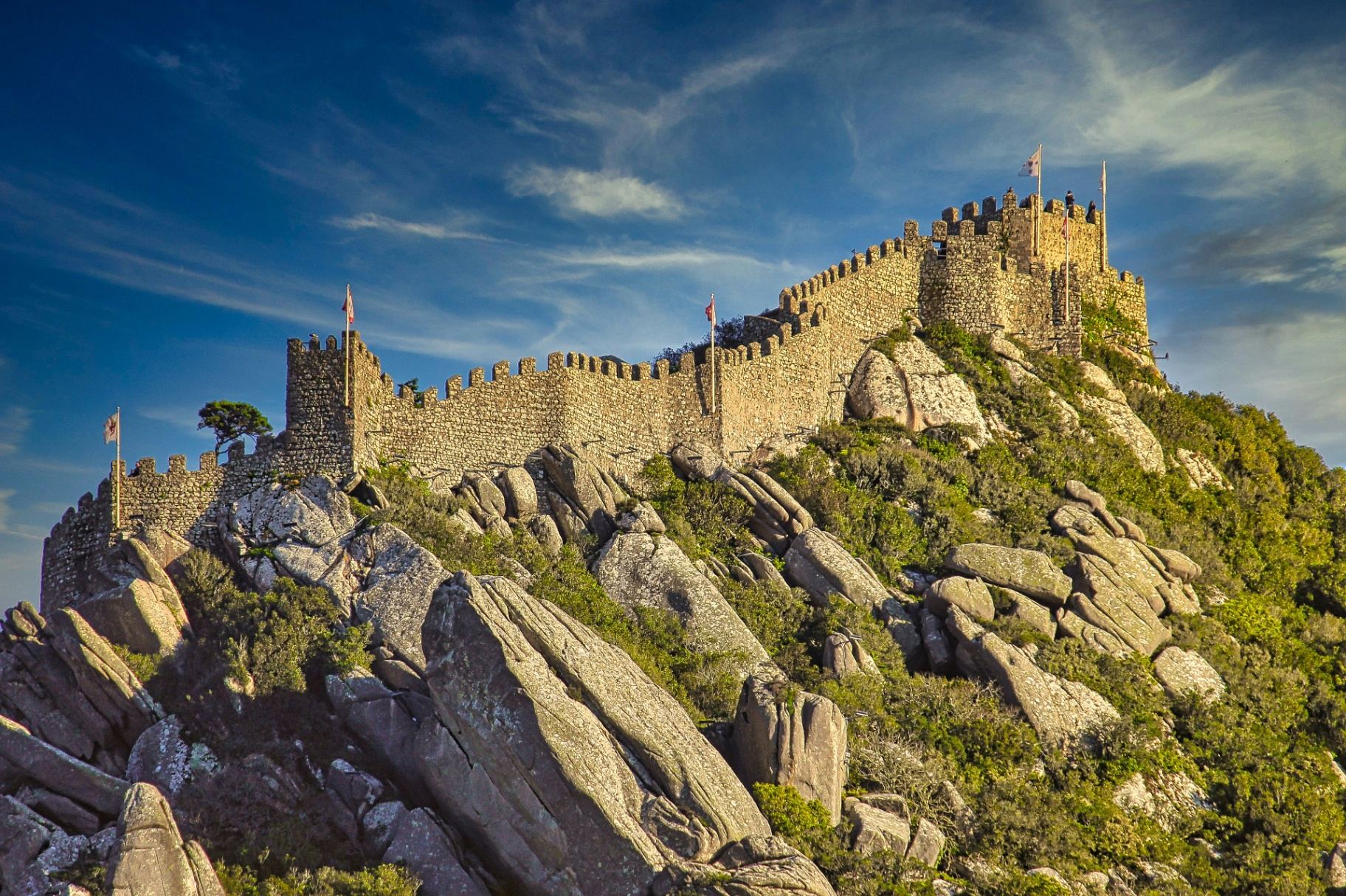 Muralhas medievais do Castelo dos Mouros sobre rochas graníticas com bandeiras ao vento e céu azul