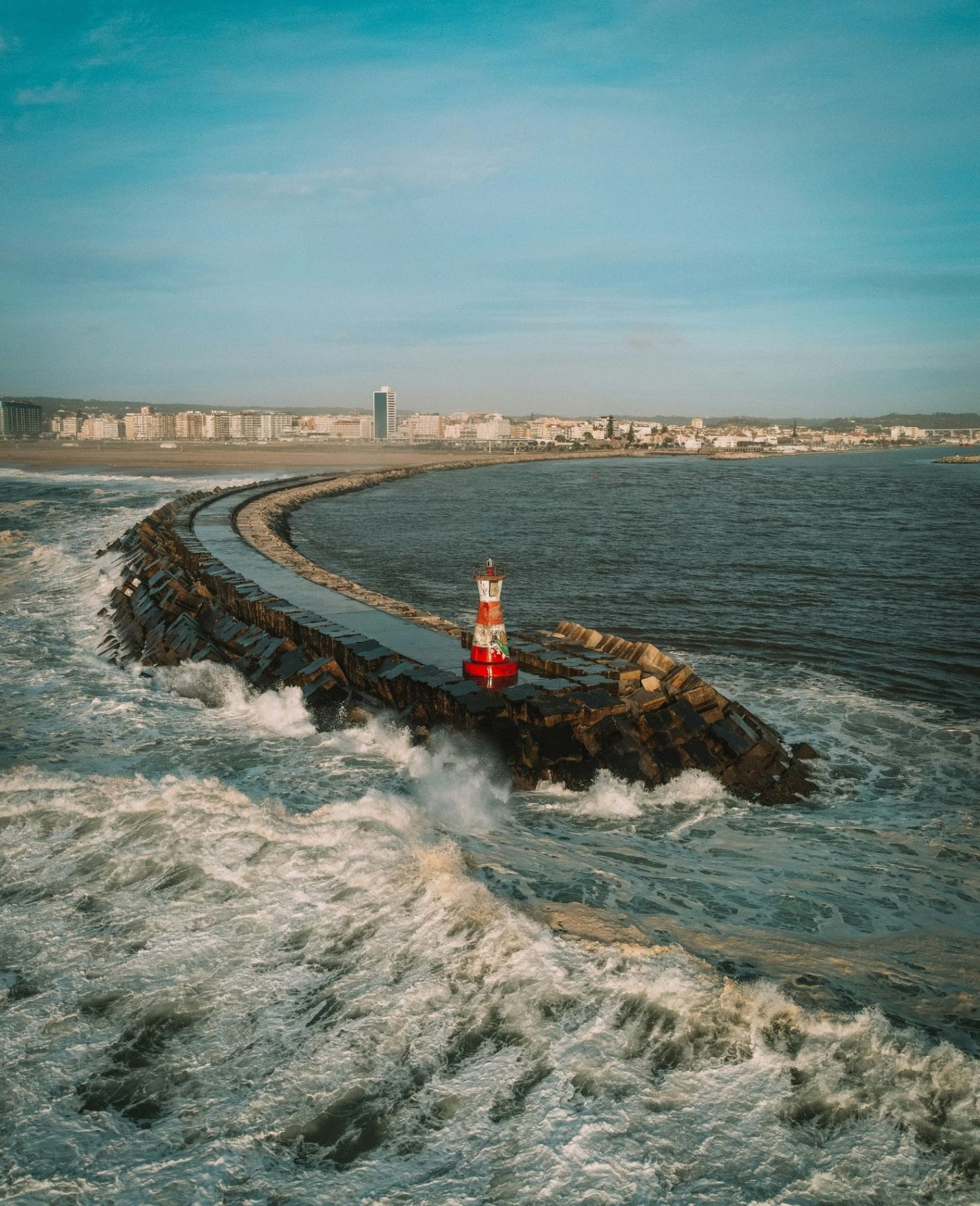 Molhe da Figueira da Foz com farol vermelho e branco rodeado por ondas do Atlântico e a cidade ao fundo