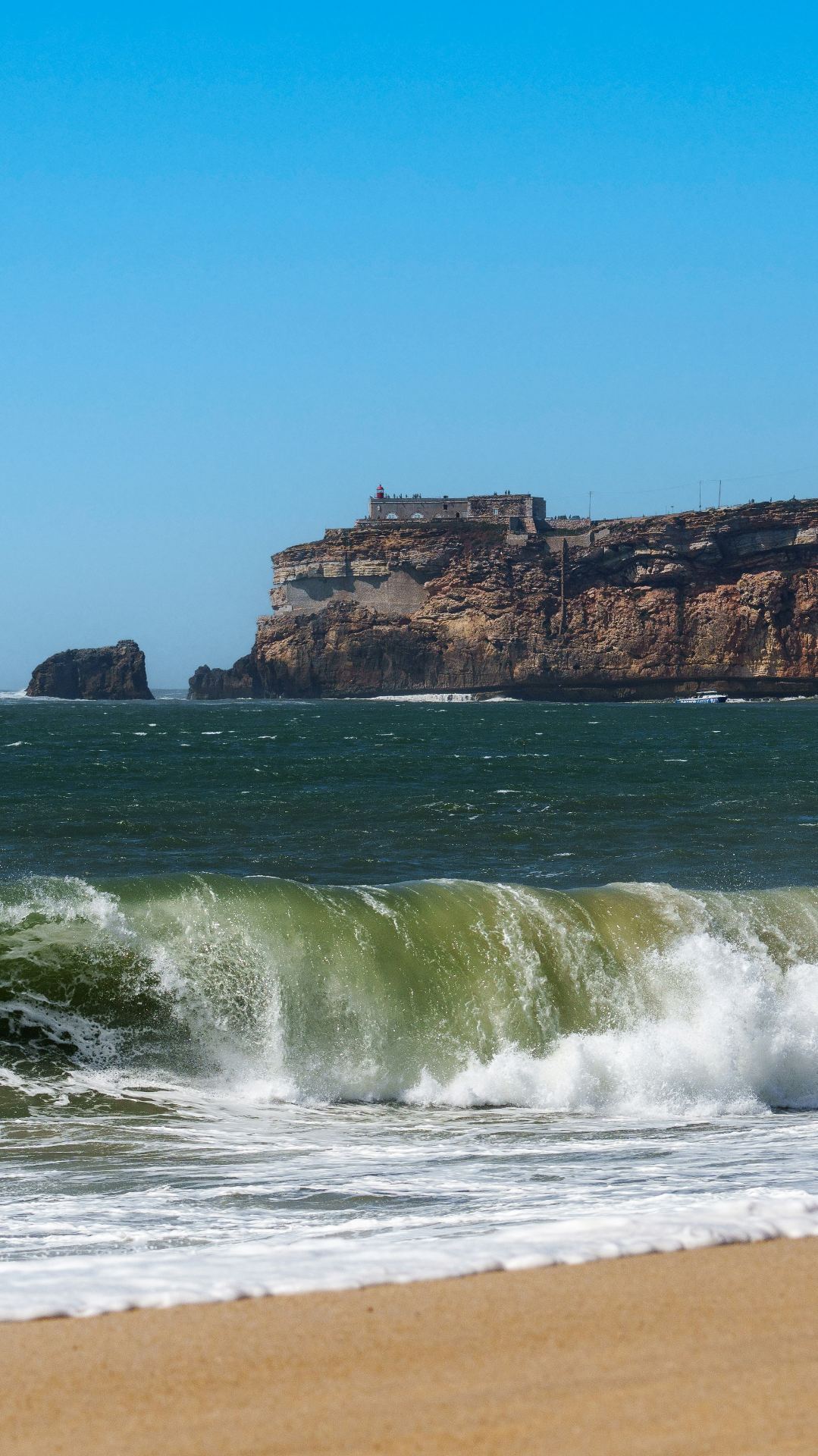 Praia da Nazaré com ondas a rebentarem e o promontório do Sítio com o Forte de São Miguel Arcanjo ao fundo
