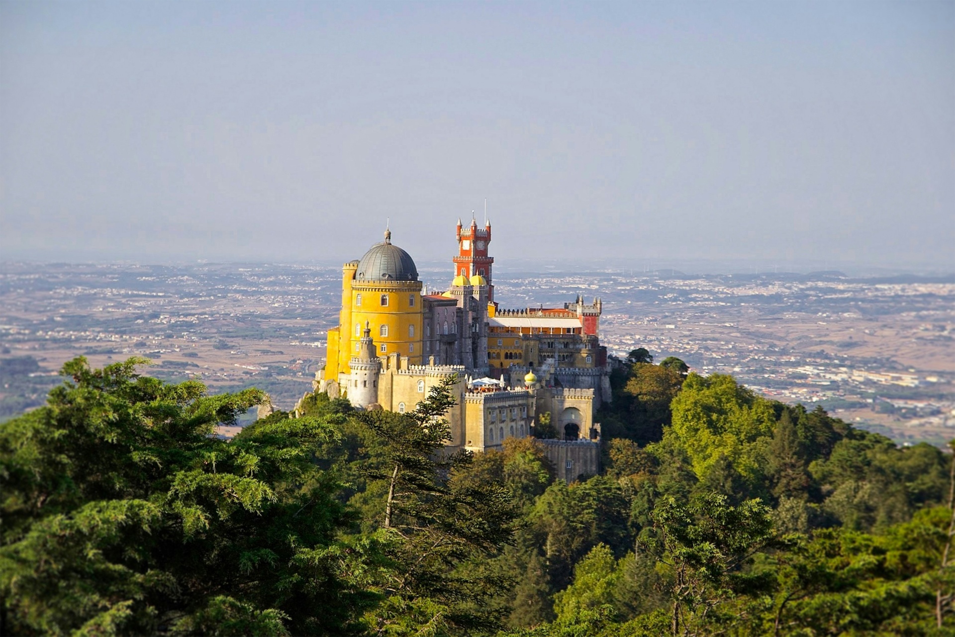 Vista panorâmica do Palácio da Pena emergindo da floresta verde da Serra de Sintra com a planície ao fundo