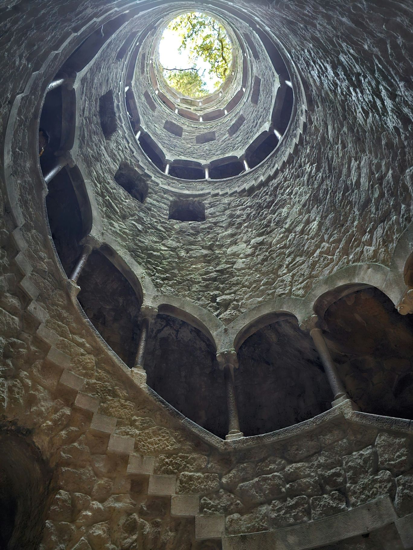 Interior do Poço Iniciático na Quinta da Regaleira visto de baixo para cima, mostrando a escadaria em espiral e a abertura circular no topo
