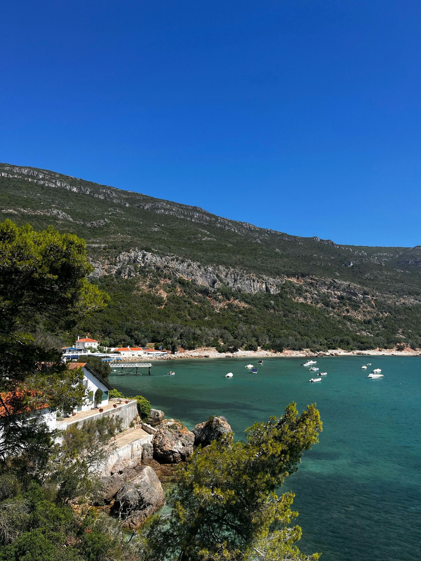 Baía de Sesimbra com águas turquesa cristalinas, barcos ancorados, casas brancas e a Serra da Arrábida ao fundo